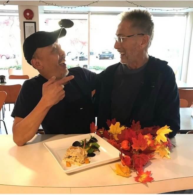 In image file autumn_gathering.jpg, two smiling men sit together at a table, with one pointing playfully at the other. A dessert and decorative autumn leaves are arranged in front of them. The scene is bright and casual, showing that they are enjoying each other’s company.